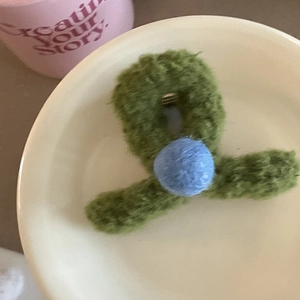 Pince à cheveux en peluche verte avec pompon bleu présentée dans un plat en céramique