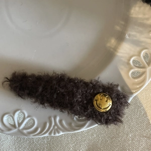 Détail d'une pince à cheveux en peluche marron doux avec un badge sourire doré sur plat