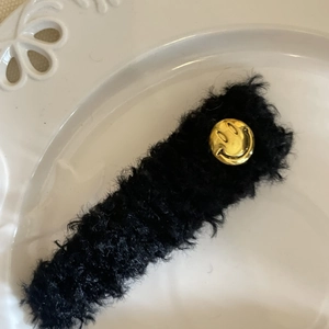 Détail d'une barrette en peluche noire avec motif smiley doré sur plateau blanc