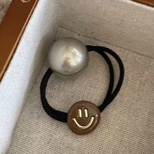 Elastic hair tie with a large glossy pearl and brown smiley face charm on beige fabric