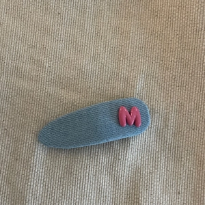 Pince à cheveux en jean bleu avec une lettre M rose sur fond beige texturé