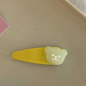 Détail d'une pince à cheveux goutte jaune avec petit ours vert sur plateau beige