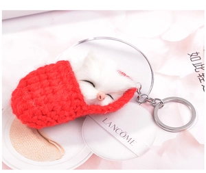 Close-up of sleeping white plush cat inside a red knitted pouch with silver keychain