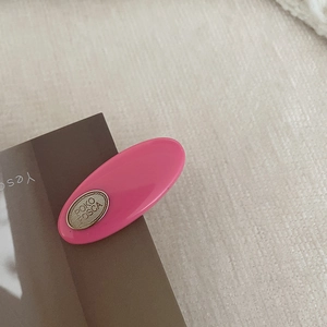 Detail shot of a rose pink oval hair clip with gold metal logo on dark paper