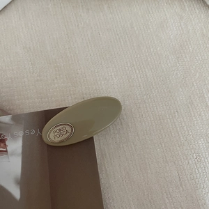 Close-up of khaki latte oval hair clip with gold metal tag on beige woven texture