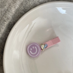 Pink metal hair clip with purple smiley face and heart-shaped rhinestone on white plate