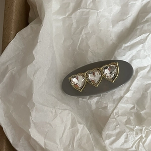 Detail of grey oval hair clip featuring three heart-shaped rhinestones and gold metal trim