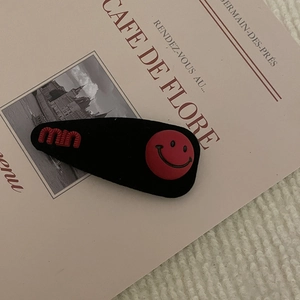 Fermaglio per capelli nero opaco con faccina rossa e scritta MU su sfondo vintage