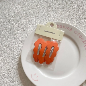 Detail showing set of three orange wavy metal hair clips in retail packaging on a plate