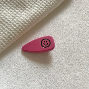 Fermaglio per capelli rosa a goccia con faccina sorridente nera su sfondo bianco