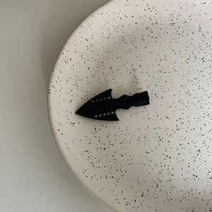 Macro detail of matte black arrowhead hair clip on a minimalist textured ceramic plate