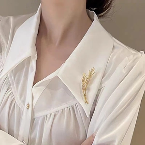 Detailed close-up of a golden wheat stalk brooch with zircons on a white silk shirt