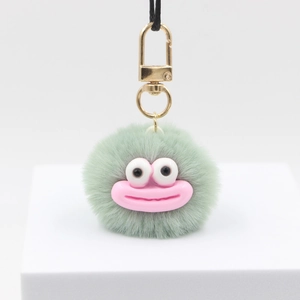Detail of green plush pompom keychain with big eyes, pink mouth and golden metal clasp