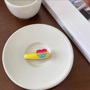 Detail of yellow hair clip with rainbow heart charm on a minimalist white ceramic plate