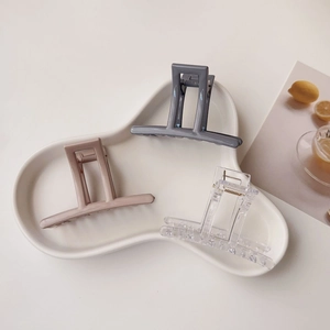 Three minimalist square acrylic hair claws in grey, pink, and clear on a ceramic tray