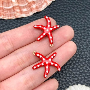 Detailed macro view of two red enamel starfish charms with gold plating held by a person