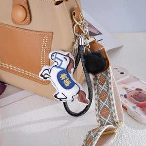 White plush horse charm with blue saddle and golden embroidery on a handbag