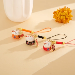 Three ceramic Maneki Neko charms with colorful woven cords and fortune tags on a warm background