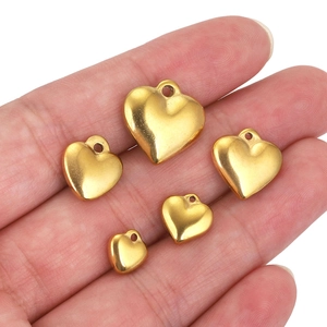 Five gold heart-shaped stainless steel pendants in various sizes held by hand on white background