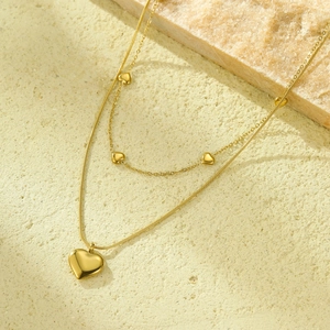 Detalle de collar de doble capa dorado con corazones y perlas sobre superficie de piedra