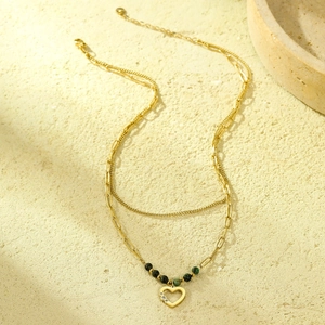 Detalle de collar doble dorado con colgante de corazón y cuentas verdes sobre piedra