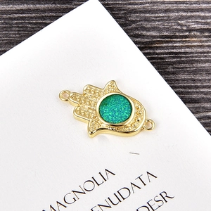 Detailed view of golden Hamsa Hand charm with green inlay on a white display card