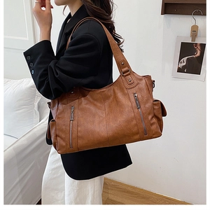 Brown leather tote bag featuring metal zipper details shown on a model in a minimalist room
