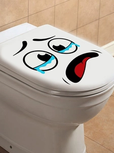 Close-up of a funny crying cartoon face sticker applied on a white toilet lid in a bathroom