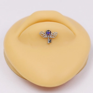 Close-up of a silver winged key lip stud with a purple gemstone on a silicone ear model