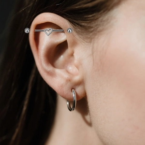 Κοντινό πλάνο βιομηχανικού piercing με κρυστάλλινη καρδιά και ασημένιο κρίκο στο αυτί
