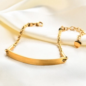 Detail view of a gold rope chain bracelet with a polished blank bar and a small heart charm