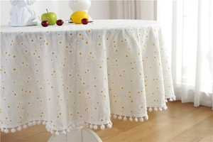 Close-up of white round cotton tablecloth with yellow daisies and white pom-pom fringe edge