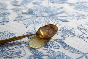 Macro shot of gold metal spoon pouring thick liquid over blue floral embroidered tablecloth