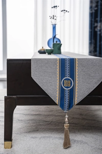 Detail of grey polyester table runner with blue embroidery and cream tassel on dark wood