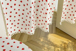 Close-up of strawberry print cotton curtain with white pom-pom trim in soft sunlight