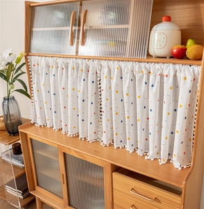 Close-up of white cotton cafe curtain with colorful polka dots and pom-pom trim on cabinet