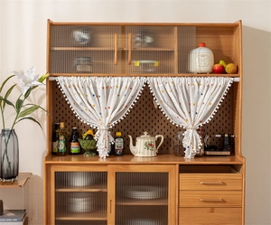 Detailed view of floral cotton curtain with white pompom trim on a kitchen cabinet