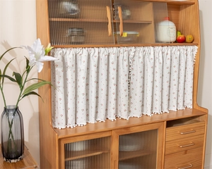 Detail of floral cotton short curtain with pom-pom trim on wooden cabinet shelf