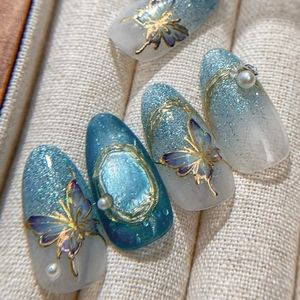 Blue gradient press-on nails with gold butterflies, glitter, and pearls on beige fabric