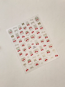 Dettaglio di adesivi per unghie con ciliegie rosse e fiocchi dorati su sfondo beige chiaro