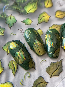 Macro view of holographic nail stickers featuring dark green and gold metallic leaves