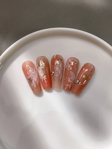 Détail de faux ongles gel pêche avec motifs floraux blancs et flocons de feuilles d'or