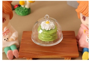 Miniature dollhouse food display with green layer cake under a clear glass cloche dome