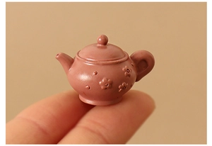 Macro shot of a miniature pink ceramic teapot with flower relief held on a fingertip