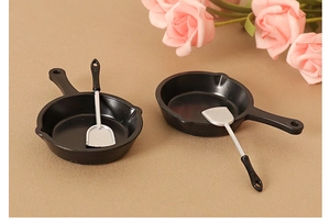 Miniature dollhouse kitchen set with black pans and silver spatulas on a neutral background