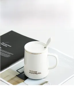 Close-up of white ceramic mug with gold rim, lid, spoon, and minimalist engraved text