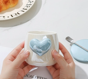 Close-up of cream ceramic heart mug with pearl glaze held by hand in a cozy cafe setting