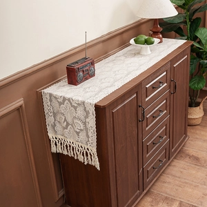 Elegant cream lace table runner with tassels on a dark wood cabinet in a cozy room