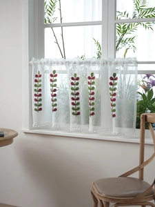 Detail of white café curtain with leaf embroidery and decorative pom-poms on the hem