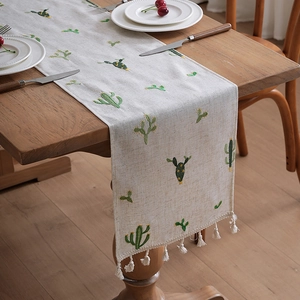 Linen table runner featuring green cactus embroidery and white tassel fringe on wood table